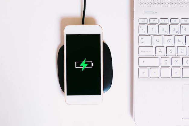 Smartphone mobile bianco con il logo di una batteria con un fulmine verde sullo schermo, in carica su una base di ricarica senza cavo accanto al laptop su un tavolo da lavoro bianco