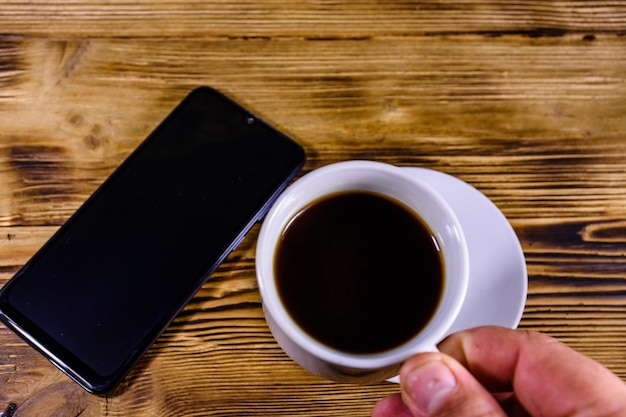 Smartphone moderno con schermo vuoto su fondo di legno e mano con una tazza di caffè