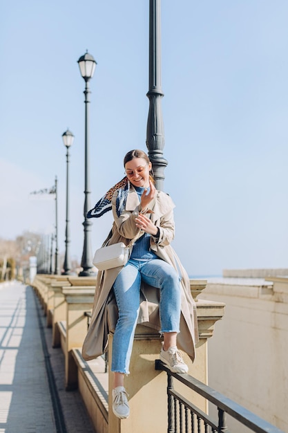Smiling brunette woman walking on the sea coast in wearing beige trench coat. Having fun lady
