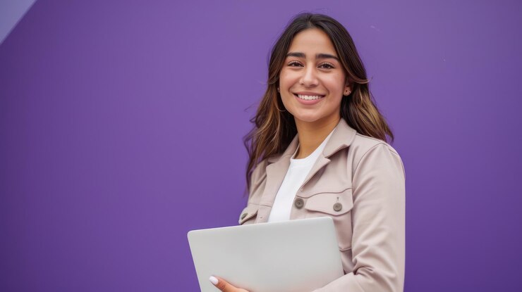 Smiling Hispanic businesswoman standing in a studio holding a laptop with a bold violet background conveying creativity and determination