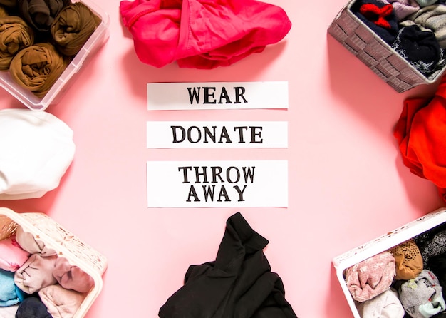 Smistamento di vestiti nell'armadio di casa per donazione, usura e scarto con note di carta su sfondo rosa tenue. Articoli di abbigliamento in scatole di tessuto e plastica.