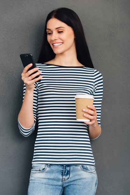 SMS ad un amico. Attraente giovane donna sorridente che tiene in mano una tazza di caffè e guarda il suo smartphone in piedi