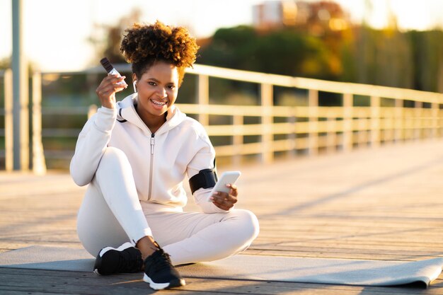 Snack fitness sorridente donna nera che mangia proteing bar dopo l'allenamento all'aperto