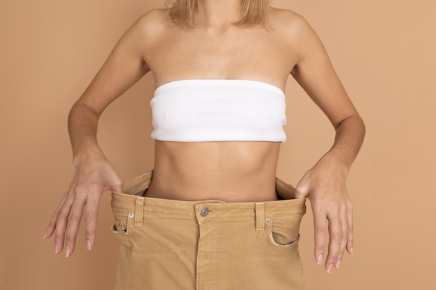 Snella signora caucasica in reggiseno bianco intimo e ampi pantaloni oversize che mostrano vita sottile su sfondo color pelle Perdita di peso Mantenere una dieta sana esercizio