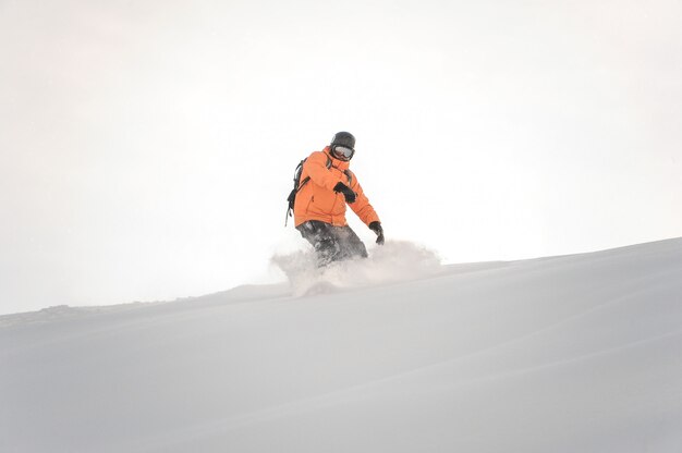 Snowboarder in abiti sportivi arancioni che guidano giù il pendio di montagna contro il cielo bianco