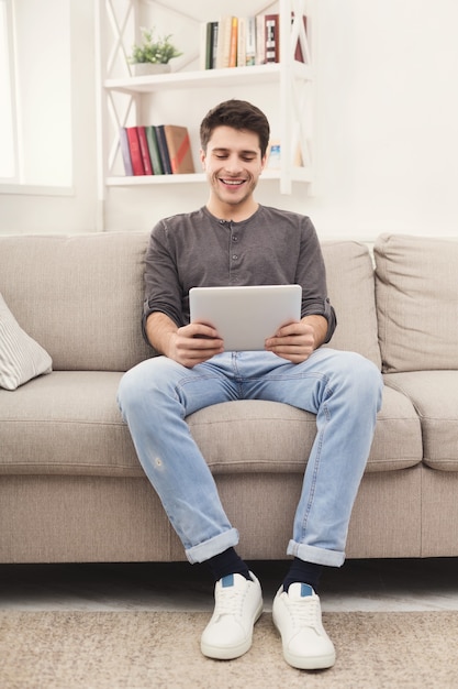 Social networks. Giovane uomo sorridente che messaggia su tablet digitale a casa, seduto comodamente sul divano beige, copia spazio