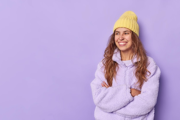 Soddisfatta bella giovane donna sta con le braccia conserte sorride con gioia mostra denti bianchi vestiti con giacca calda e cappello isolato su sfondo viola spazio vuoto per i tuoi contenuti pubblicitari