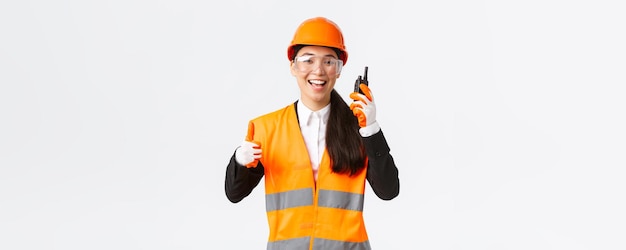 Soddisfatto tecnico industriale ingegnere femminile asiatico sorridente in casco di sicurezza e uniforme che mostra pollice in su mentre loda l'ottimo lavoro usando il walkietalkie dà il permesso di lavorare