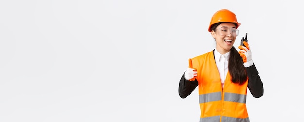 Soddisfatto tecnico industriale ingegnere femminile asiatico sorridente in casco di sicurezza e uniforme che mostra pollice in su mentre loda l'ottimo lavoro usando il walkietalkie dà il permesso di lavorare