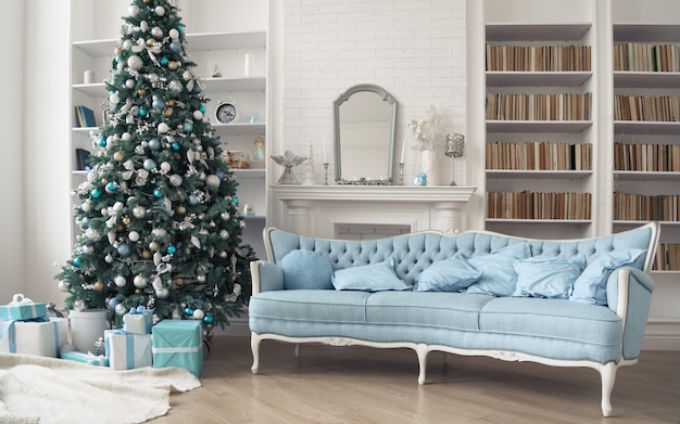 Sofà blu francese classico e albero di Natale decorato con i contenitori di regalo sotto in salone con gli scaffali di libro