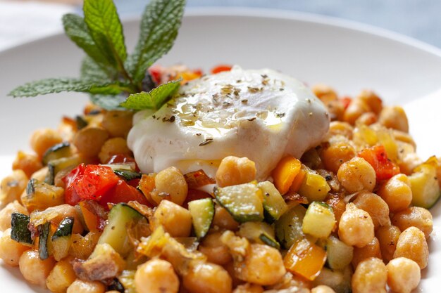 Soffriggere vegetariano piccante fatto in casa con ceci e dadi di pepe e zucchine con un uovo fritto e qualche decorazione di menta in un piatto bianco. Avvicinamento.