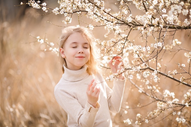 Sognando una ragazza bionda con gli occhi chiusi nel giardino fiorito vicino ai fiori bianchi di sakura. Ciliegio primaverile e caldo tema soleggiato