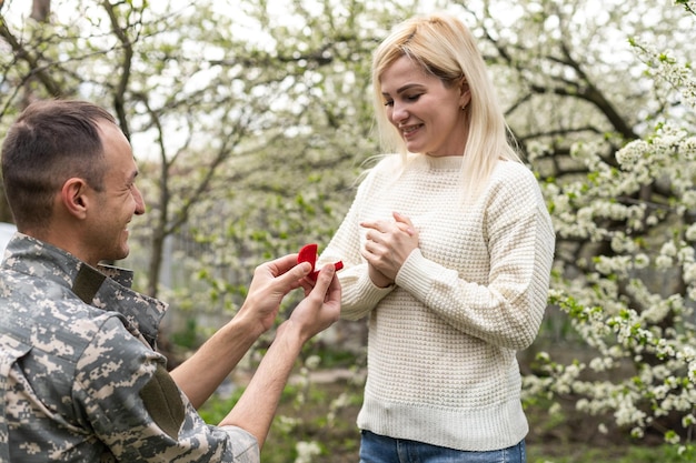 soldato in ginocchio che chiede il matrimonio tenendo le mani della sua compagna e donando un anello di fidanzamento. Concetto di patriottismo, guerra, soldato.