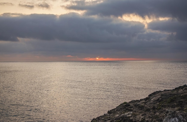 Sole rosso che sorge sopra il mare dietro le nuvole, costa rocciosa vicino