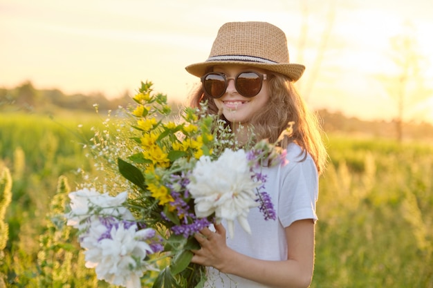 Soleggiato ritratto all'aperto della bambina di 9, 10 anni in occhiali da sole cappello con bouquet di fiori in prato