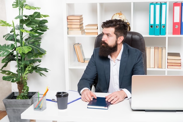 Solita giornata in ufficio. Il capo dei pantaloni a vita bassa barbuto dell'uomo si siede nell'interiore dell'ufficio della poltrona di pelle. Capo sul posto di lavoro. Responsabile abbigliamento formale stile aziendale funzionante. Concetto di uomini d'affari. Capo impegnato in ufficio.