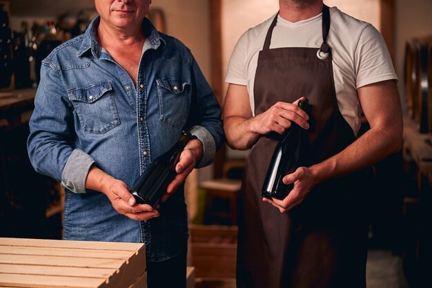 sommelier e il suo studente in piedi con bottiglie di vino senza etichetta in una cantina