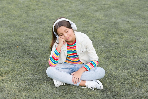 Sonnecchia goditi la primavera natura all'aperto bambino studio audiolibro nuova tecnologia bambino ascolta musica sull'erba verde stile di vita sano e relax yoga ragazza bambino piccolo in cuffia playlist estiva