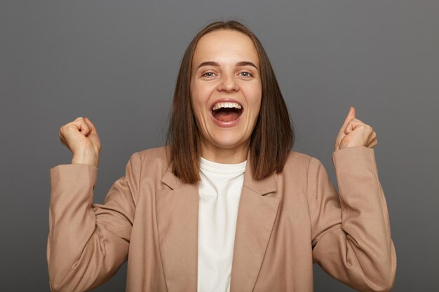 Sono la campionessa Donna entusiasta motivata con i capelli scuri che indossa una giacca in piedi contro il muro grigio urlando di gioia e alzando i pugni celebrando con successo una vittoria incredibile