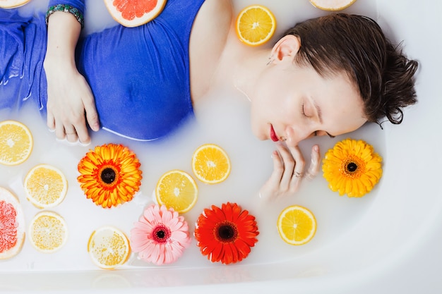 Sopra il colpo di donna in abito blu che giace nella vasca da bagno piena di acqua bianca con fiori e agrumi