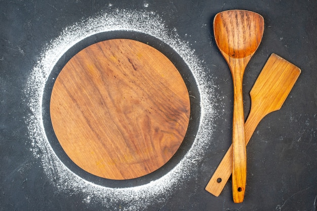 Sopra la vista del tagliere rotondo con farina intorno a cucchiai di legno su sfondo di colore scuro con spazio libero