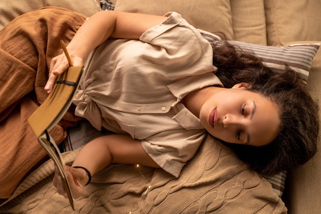 Sopra la vista della donna di razza mista sognante calma sdraiata sul letto e leggere il libro