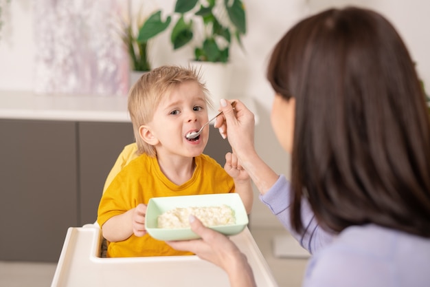 Sopra la vista della spalla della madre che alimenta il figlio con il porridge usando il cucchiaio in cucina