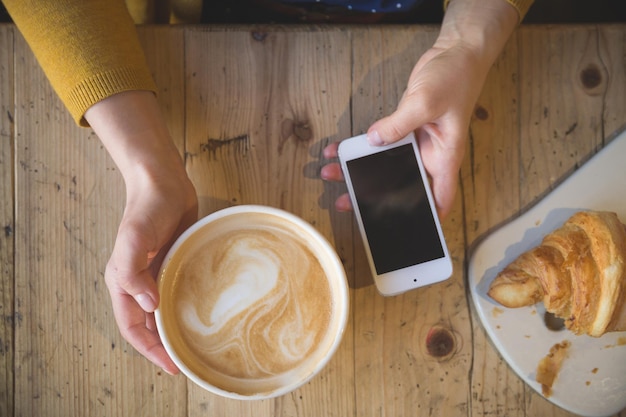 Sopra la vista delle mani femminili che tengono una tazza di caffè calda e con lo smartphone sul tavolo di legno