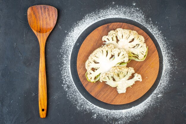 Sopra vista di cavolfiori freschi su tagliere rotondo e cucchiaio di legno su superficie di colore scuro