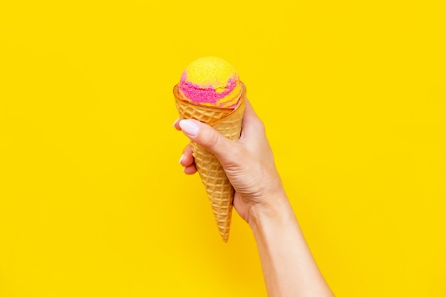 Sorbetto di gelato alla frutta rosa gialla dolce in un cono di cialda con sapore di banana e fragola in una mano femminile isolata su una parete gialla di colore brillante