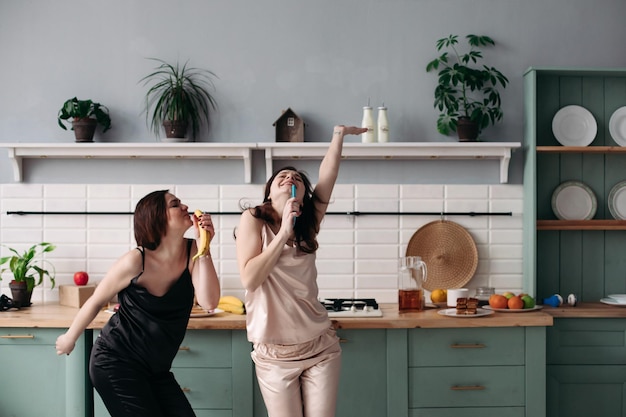 Sorelle attraenti in pigiama bianco e nero che ascoltano musica e ballano in cucina al mattino Belle ragazze felici che si divertono mentre cucinano la colazione Amici castani che cantano e saltano