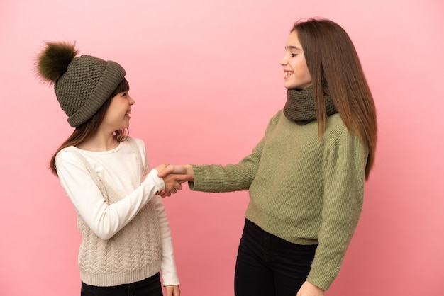 Sorelline che indossano abiti invernali isolati su sfondo rosa che si stringono la mano dopo un buon affare