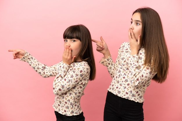 Sorelline ragazze isolate su sfondo rosa che puntano il dito sul lato con una faccia sorpresa