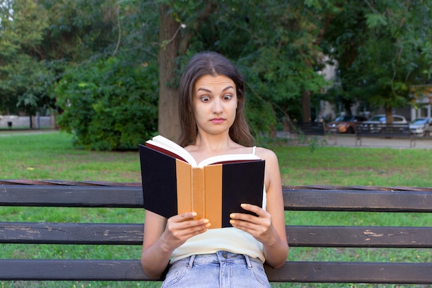 Sorpresa confusa e con gli occhi spalancati, la donna sta leggendo un libro sulla panchina del parco.