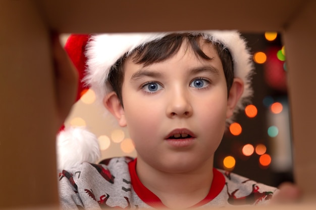 sorpresa di emozione in un ragazzo con un maglione di Capodanno e un cappello di Babbo Natale guarda in una scatola con un regalo, una vista dall'interno, su uno sfondo