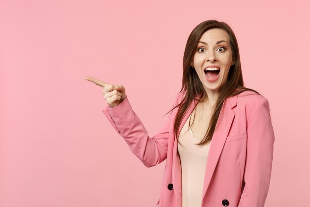 Sorpresa divertente giovane donna che indossa una giacca tenendo la bocca aperta, puntando il dito indice da parte isolato su sfondo rosa pastello in studio. Persone sincere emozioni, concetto di stile di vita. Mock up spazio di copia.