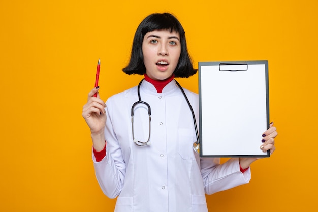 Sorpresa giovane bella ragazza caucasica in uniforme da medico con stetoscopio che tiene penna e appunti