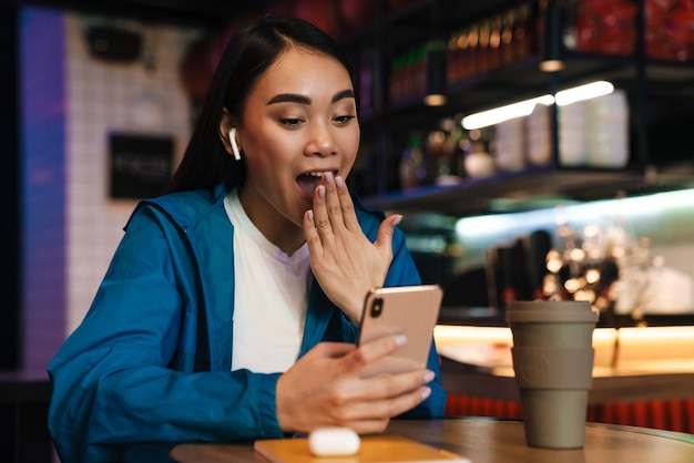 sorpresa giovane donna asiatica che usa il telefono cellulare e gli auricolari wireless mentre è seduta al bar