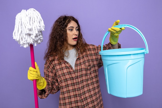 Sorpresa giovane donna delle pulizie che indossa guanti che tengono mop guardando il secchio in mano