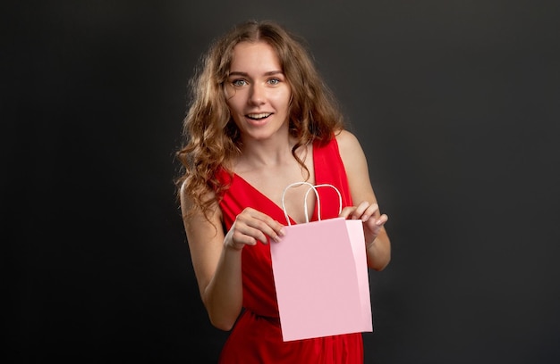 Sorpresa per le vacanze Regalo di compleanno Donna stupita in abito rosso felice di ricevere un regalo in una borsa della spesa rosa mockup di marca isolata su sfondo scuro dello spazio della copia