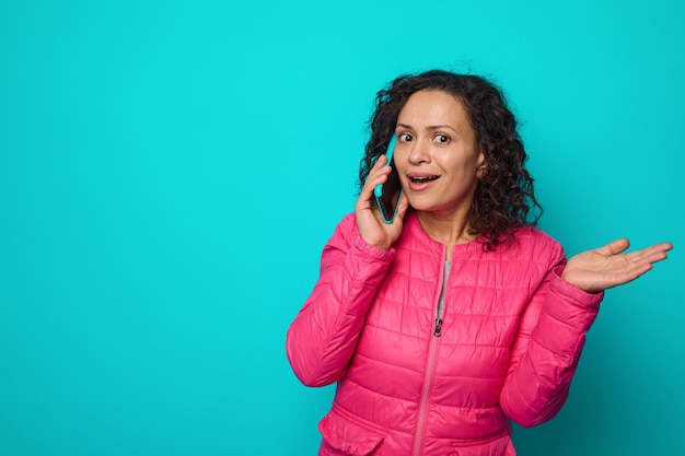 Sorpresa scioccata emotiva dai capelli ricci bella donna in giacca rosa brillante parla al telefono cellulare e gesti con il palmo della mano in alto, isolato su sfondo colorato blu con lo spazio della copia