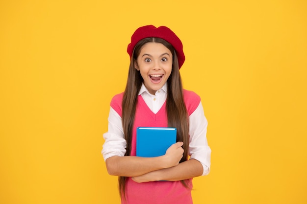 Sorpresa studentessa adolescente in berretto francese tenere quaderno su sfondo giallo scuola