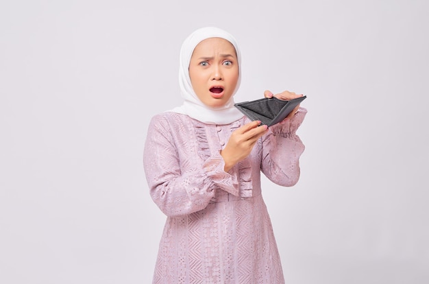 Sorpreso bella giovane donna musulmana asiatica che indossa l'hijab e abito viola che mostra portafoglio vuoto e guardando la fotocamera isolata su sfondo bianco studio