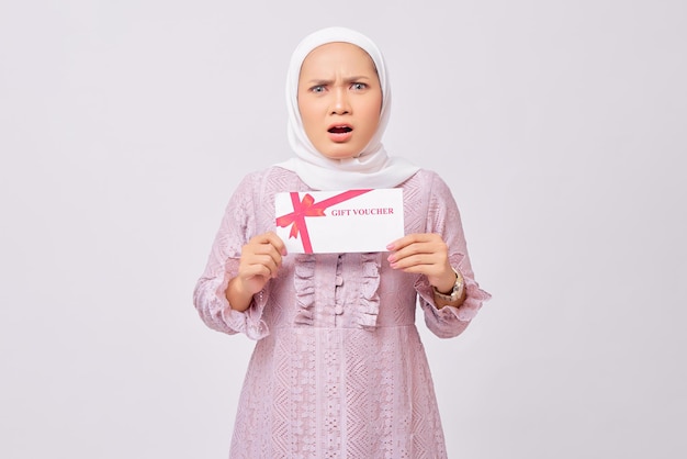 Sorpreso bella giovane donna musulmana asiatica che indossa l'hijab e vestito viola tenendo il certificato del buono regalo e guardando la fotocamera isolata su sfondo bianco studio