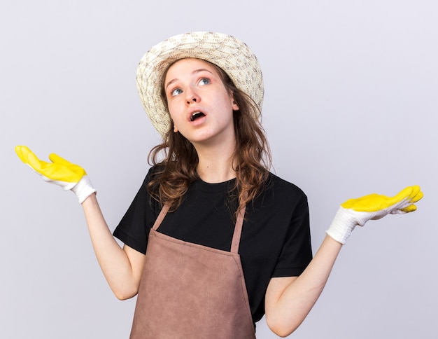 Sorpreso giovane giardiniere che indossa un cappello da giardinaggio con guanti che allargano le mani isolate sul muro bianco