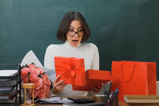 sorpreso giovane insegnante femminile che tiene e guarda il presente seduto alla scrivania con gli strumenti della scuola in classe