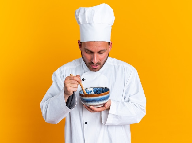 Sorpreso giovane maschio caucasico cuoco in uniforme da chef e cappuccio che tiene ciotola e cucchiaio di legno al suo interno guardando dentro la ciotola