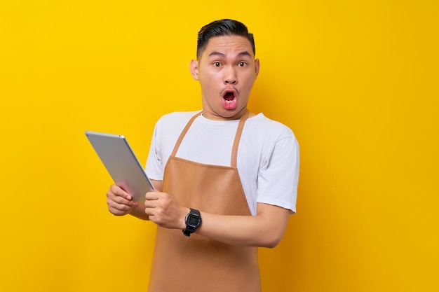 Sorpreso giovane uomo asiatico barista barman dipendente che indossa grembiule marrone lavora in caffetteria tenendo tablet digitale e guardando la fotocamera con la bocca aperta su sfondo giallo Avvio di piccole imprese