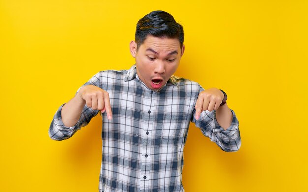 Sorpreso giovane uomo asiatico bello che indossa una camicia a quadri che punta le dita verso il basso invitando i clienti a un evento speciale isolato su sfondo giallo Concetto di stile di vita delle persone