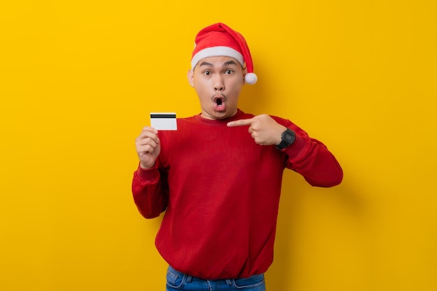 Sorpreso giovane uomo asiatico in cappello di Babbo Natale che punta il dito contro la carta di credito su sfondo giallo studio celebrazione vacanze di Natale e concetto di Capodanno